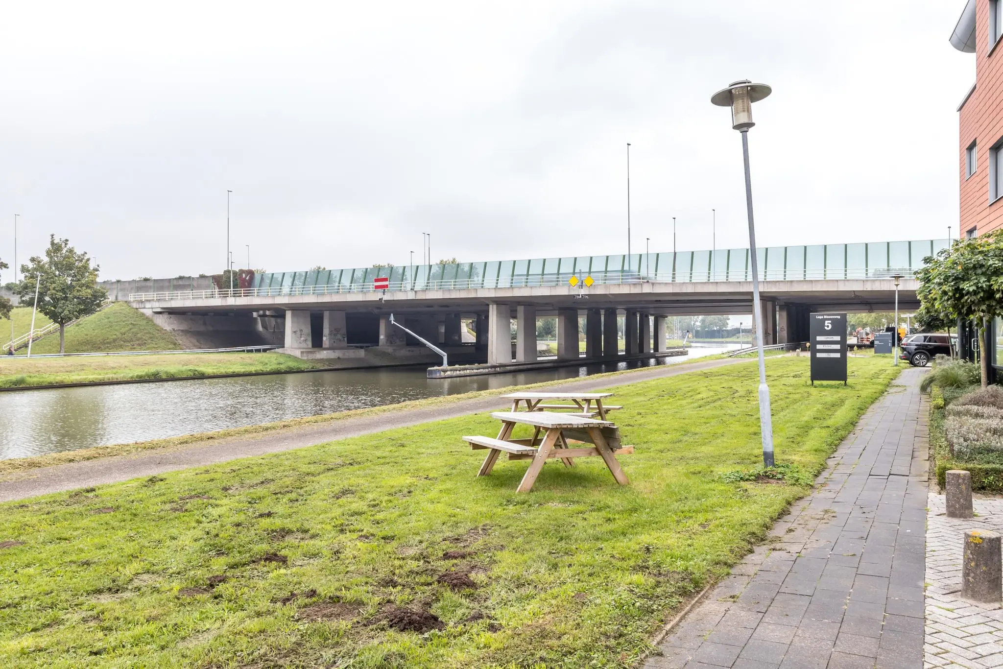 Picknicktafel op een grasveld langs het water bij de Lage Biezenweg met zicht op een brug en kantoorpand.
